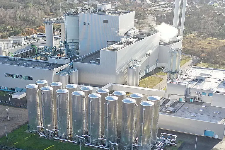 Luftaufnahme einer großen Industrieanlage mit mehreren silbernen Silos, umgeben von Wald